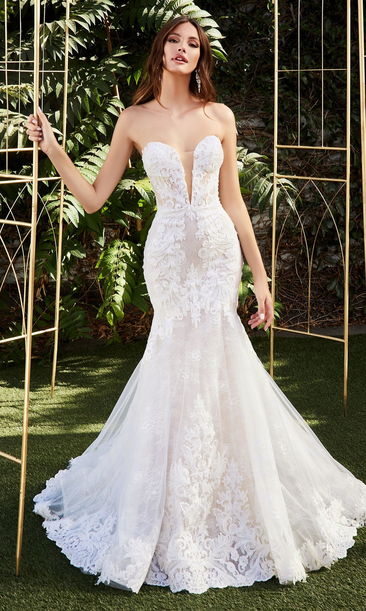A woman wearing the Ladivine CD928 Strapless Mermaid Wedding Gown with a deep V neckline stands outdoors on green grass, surrounded by gold metal arches and leafy plants.